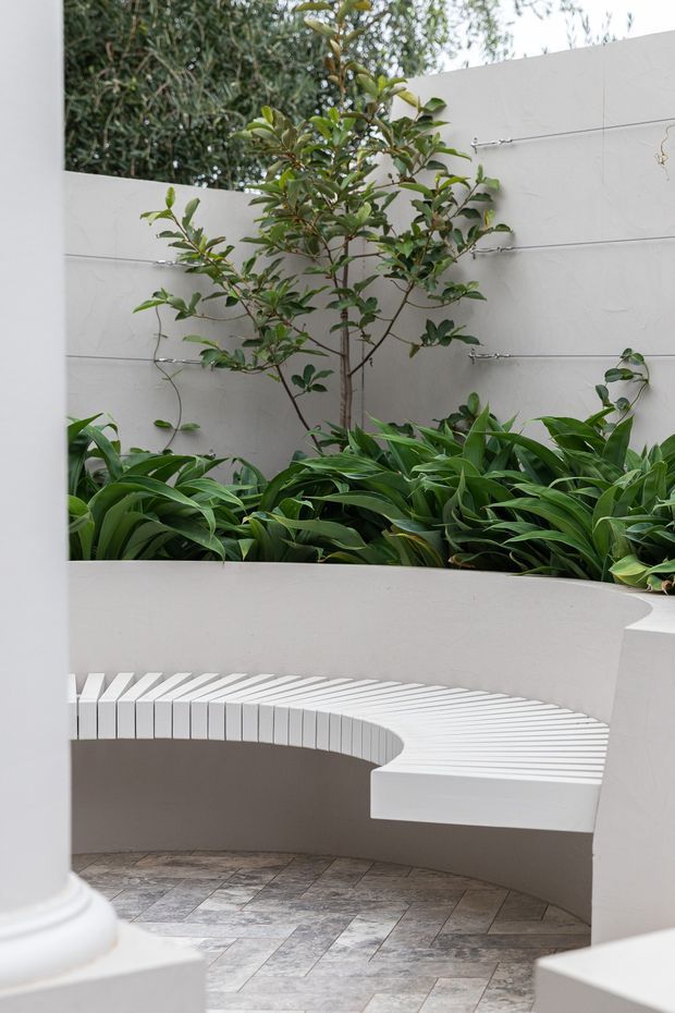 A serene landscaped courtyard with a curved built-in white bench wrapping around a planted garden bed. White rendered walls and classical column details frame the space, while grey stone-look paving and dense green foliage create a calm, minimalist outdoor retreat.