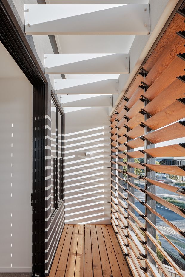 A bright, narrow covered balcony or terrace with timber decking, black-framed glazing, and a prominent timber louvred screen along one side. White overhead beams and strong sunlight create repeated linear shadows across the walls and floor. The space has a contemporary, structured architectural character.