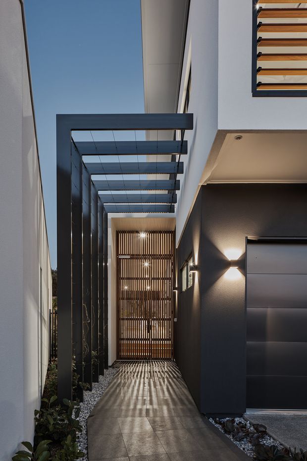 A narrow modern external passage beside a house, leading toward a timber slatted gate or screening element. Dark steel posts support a projecting overhead pergola with translucent or open panels, creating strong linear shadows across large-format tiled paving. White and charcoal rendered walls, integrated wall lights, gravel garden beds, and a dark sectional garage door frame the walkway. The view emphasizes a refined, contemporary architectural connection between the garage and rear or side entry.
