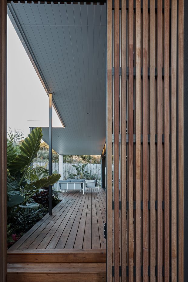 A narrow covered outdoor deck or entry terrace beside a modern home. The foreground is dominated by a tall vertical timber slat screen or gate with dark metal framing. Timber decking and steps lead toward a small seating area, beneath a cool grey-blue soffit supported by slim dark posts. Lush tropical planting borders the deck, and a pale privacy fence is visible in the background. The composition emphasises layered timber, screening, and indoor-outdoor connection.