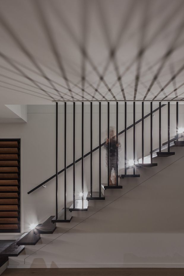 Interior view of a modern staircase with open-riser timber treads, slim black vertical balusters, and a minimalist black handrail. The stair rises diagonally across a light neutral wall, with integrated lighting beneath several steps and a large reflective ceiling surface creating repeated linear reflections. A dark timber-clad wall or doorway is visible at left.