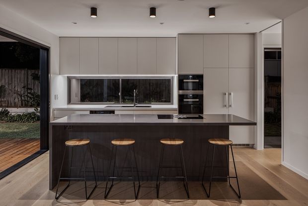 Contemporary kitchen with a large dark wood island and four slim bar stools, pale full-height cabinetry, integrated appliances, a long horizontal window above the sink, and light timber flooring. Dark-framed glazing opens toward an outdoor area, while recessed and cylindrical pendant lighting provide a clean, refined fit-out.
