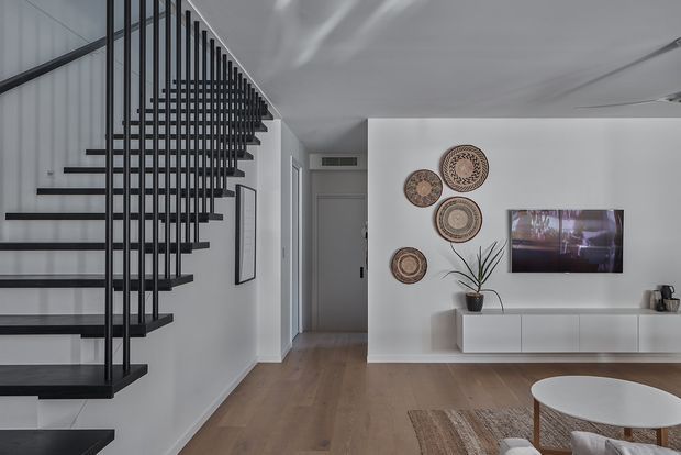 A contemporary open-plan living area with a floating black staircase and vertical metal balustrade dominating the left side. The room has white walls and ceiling, warm timber flooring, a minimalist white TV console, wall-mounted television, woven circular wall decor, and a partially visible round coffee table and rug. A hallway and doorway extend beyond the living space.