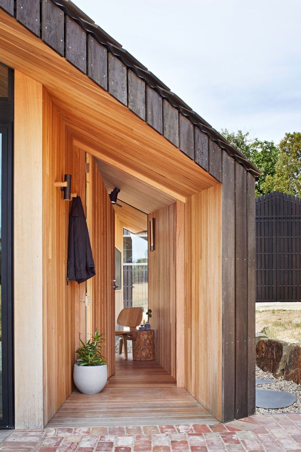 A close view of a covered residential entrance porch and recessed front door, framed in warm vertical timber lining. The entry connects visually to an interior hallway or room beyond, with a small chair and side table visible inside. Dark weathered cladding and a dark vertical-slat boundary fence contrast with the timber, while a red-toned brick paving surface forms the threshold.