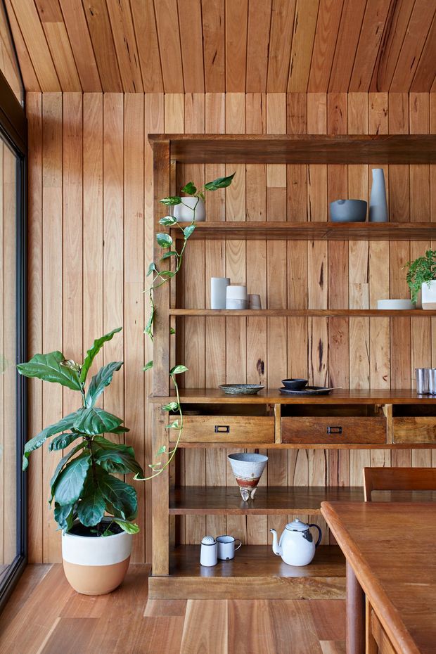 Warm, richly timber-lined dining area featuring a full-height built-in open shelving and drawer unit displaying ceramics and tableware. A large potted plant adds greenery beside the joinery, while a timber dining table and chair are visible in the foreground. The walls, ceiling, shelving, cabinetry, and flooring are all dominated by natural wood, creating a cohesive, intimate interior.