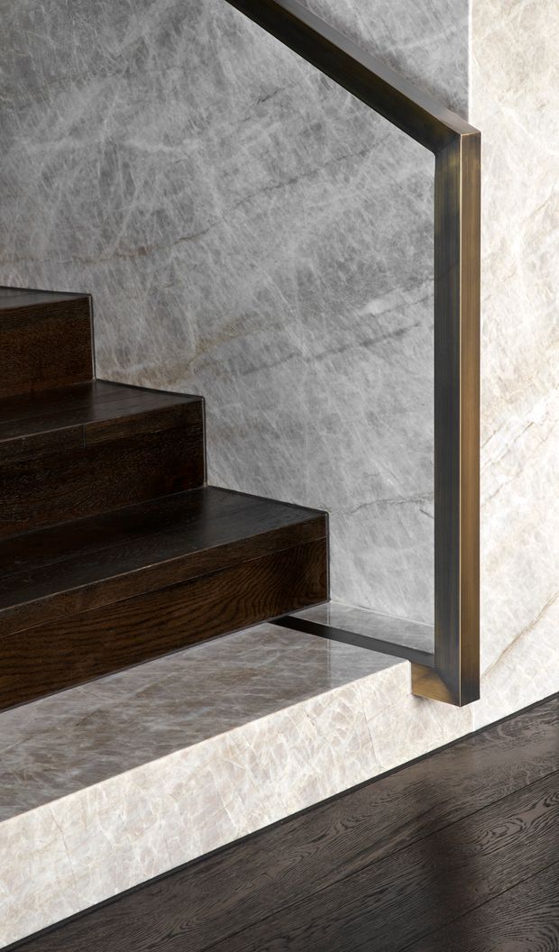 Close-up architectural detail of a contemporary stairway with dark timber treads and flooring, pale veined stone wall and riser surfaces, and a slim dark metal handrail with a square vertical support. The composition highlights contrasting natural materials and refined detailing.