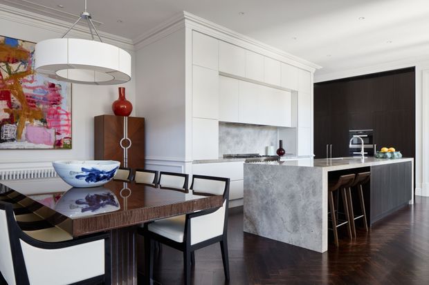 A refined open-plan dining and kitchen interior with white paneled cabinetry, a large grey stone island, dark timber flooring, and a formal dining table with upholstered black-framed chairs. The space features decorative wall mouldings, a sculptural pendant light, contemporary artwork, and a dark feature wall in the kitchen.