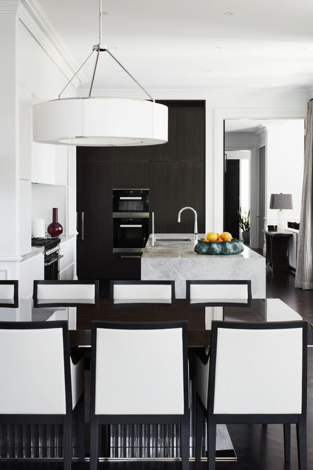 A refined open-plan kitchen and dining area with a dramatic black built-in kitchen, pale stone island benchtop, white walls and detailed ceiling mouldings. A large white drum pendant hangs above the island, while a dark dining table with white upholstered chairs occupies the foreground. The kitchen includes integrated ovens, a sink with a curved chrome tap, and dark timber cabinetry. A living or office area is visible through the doorway at right.