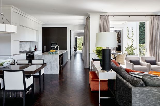 A refined open-plan interior combining a contemporary kitchen, dining area, and living room. The space features white built-in cabinetry, a pale stone island, dark timber flooring, tailored upholstered furniture, full-height curtains, and classic wall and ceiling mouldings. Large windows bring in abundant natural light, while dark accents and warm orange upholstery add contrast.