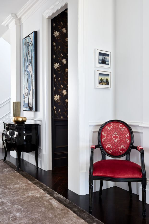 A refined residential hallway or entry area with white painted walls, ornate architectural mouldings and wainscoting, dark timber flooring, and a large neutral rug. A glossy black console table with a brass bowl sits beneath framed artwork, while a richly upholstered red and black accent chair is positioned against the wall. An open doorway reveals dark floral wallpaper and black panelled detailing, reinforcing the formal, decorative character of the space.