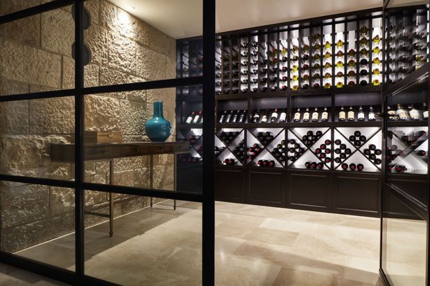 A luxurious, dimly lit wine storage room viewed through black-framed glass doors. The room features extensive dark built-in wine cabinetry and geometric bottle racks, a pale stone or tile floor, textured natural stone walls, and a slim console table with a blue decorative vase. Integrated lighting highlights the wine collection and shelving.
