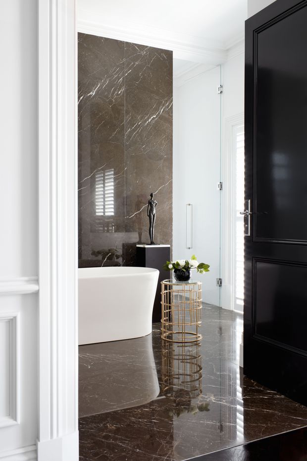A refined, luxurious bathroom viewed through an open black paneled door. The room features a freestanding white bathtub, floor-to-ceiling dark brown veined marble wall and floor surfaces, a clear glass shower enclosure, white decorative wall mouldings, and reflective polished finishes. A small gold metal side table with flowers and a sculptural black pedestal accent the space.