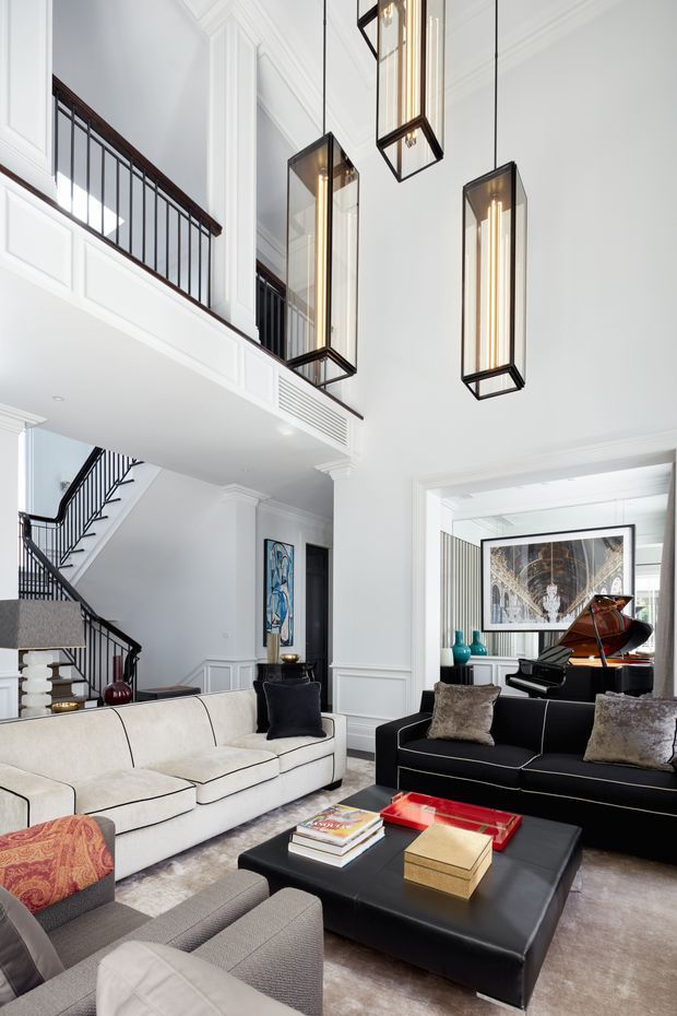 Bright, double-height formal living room with a mezzanine balcony and staircase, white paneled walls and elaborate ceiling mouldings, dark metal balustrades, and several suspended rectangular pendant lights. The room is furnished with contrasting white and black sofas, grey armchairs, a large black coffee table, and a grand piano beneath framed artwork. The space has a refined, high-contrast traditional-contemporary character.