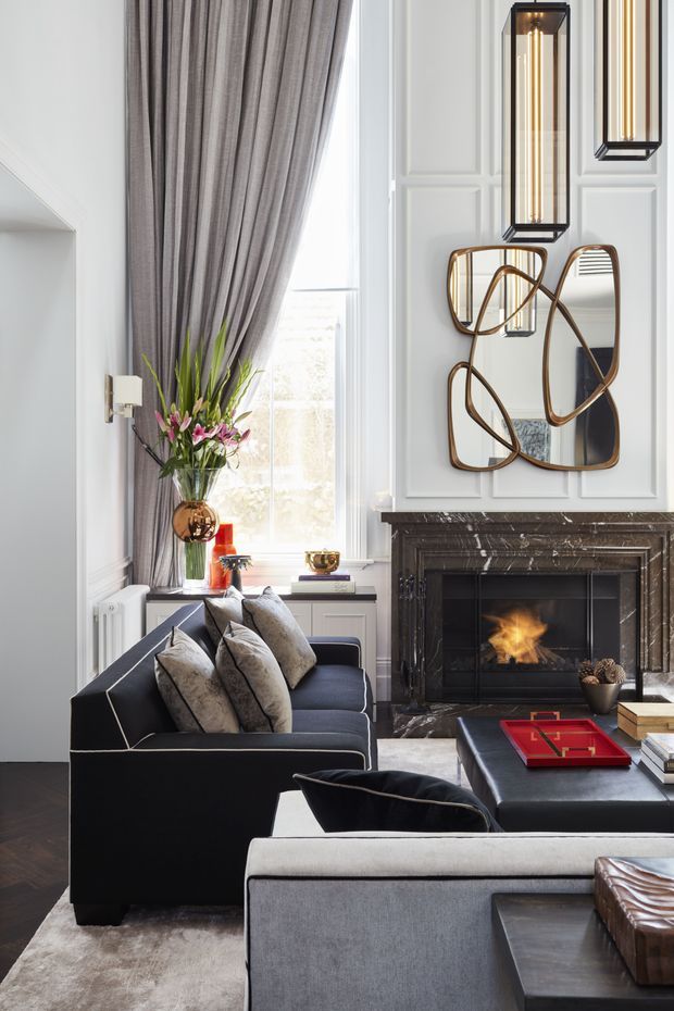 Elegant formal living room with a dark upholstered sofa and armchairs arranged around a low coffee table, a marble fireplace with an active fire, tall grey curtains, white decorative wall panelling, sculptural wall mirror, pendant lighting, and floral styling. The room has a refined, layered and luxurious appearance.