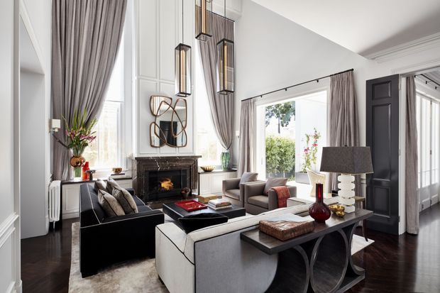Elegant, bright formal living room with tall ceilings, dark timber flooring, white decorative wall panelling, a traditional fireplace, floor-length taupe curtains, and large windows. The room is furnished with sofas, armchairs, a central coffee table, and a console table, with sculptural pendant lights and restrained contemporary decor. A dark paneled doorway opens toward an adjacent space.