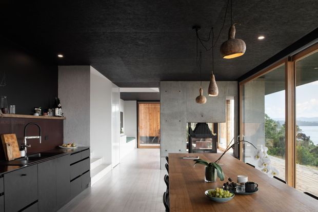 A dark, sophisticated open-plan kitchen and dining area with a long timber dining table beside full-height windows overlooking a natural landscape and water. The kitchen features matte black cabinetry, a dark benchtop, an integrated sink, and open shelving. The space has a textured dark ceiling, exposed concrete feature walls, warm timber doors and window frames, and sculptural pendant lights. A glimpse of a bathroom or adjacent room is visible through the hallway, but the kitchen-dining zone dominates.