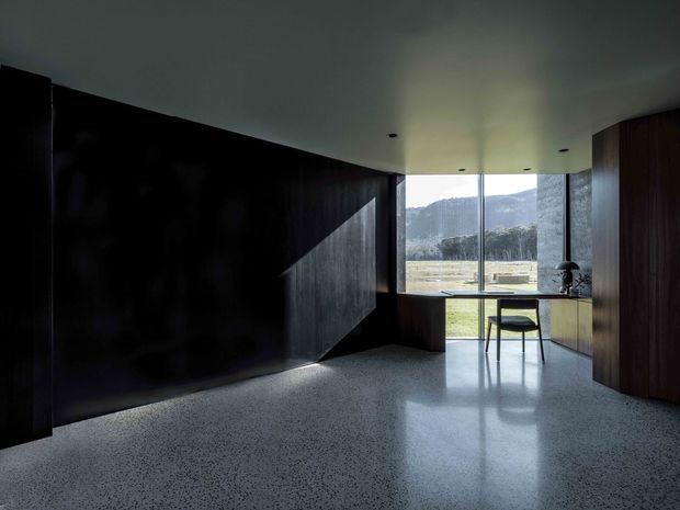 Dark, minimalist interior workspace or study with extensive dark timber wall panelling and built-in joinery, a compact desk facing full-height glazing, and a single chair. The polished speckled floor and pale ceiling contrast with the deep timber surfaces. The glazing provides a view toward an open rural landscape and mountains, while daylight creates a strong geometric shadow across the wall.
