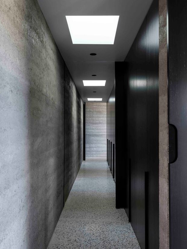 A narrow, long modern hallway with a minimalist, dark-toned fit-out. The corridor features a textured raw concrete wall on the left, full-height black or very dark timber joinery and doors on the right, and a speckled polished terrazzo-style floor. Recessed rectangular ceiling lights create a strong rhythmic perspective, while a textured concrete wall closes the view at the far end.