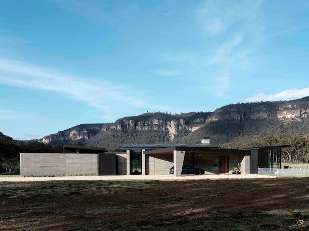 A low, horizontally composed modern residence set against a dramatic wooded escarpment. The house features flat roof planes, exposed concrete walls, dark vertical cladding, extensive glazing, and an integrated open carport. The foreground is an undeveloped landscape, while the building sits prominently within a rural or bush setting.