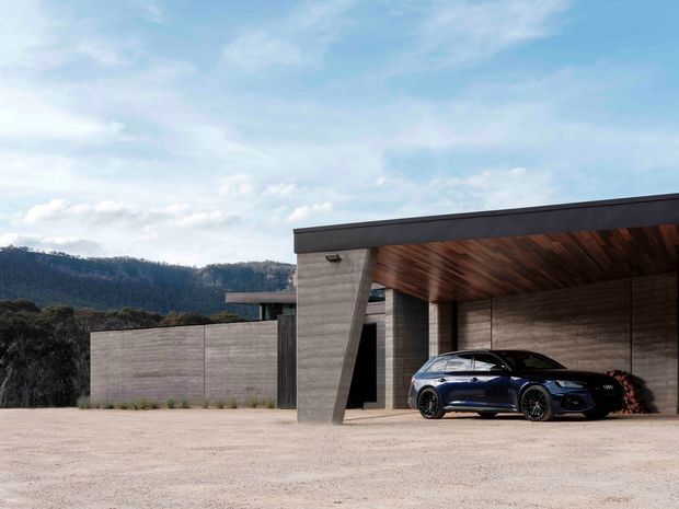 Modern single-storey concrete residence with a broad flat-roofed carport or garage canopy. The structure features exposed board-formed concrete walls and a warm timber-lined soffit, with a dark-framed opening and a vehicle parked beneath the shelter. A pale gravel forecourt and wooded hills are visible beyond the property.