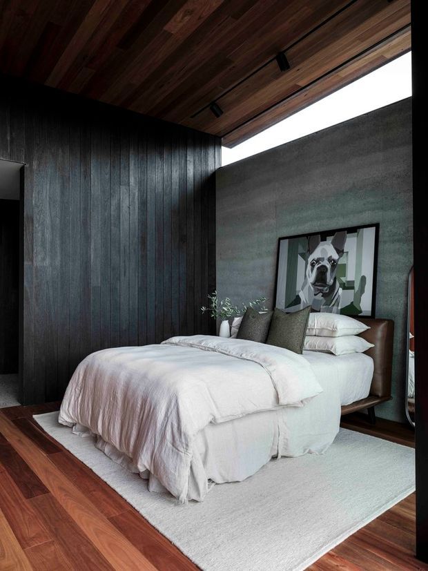 A moody, sophisticated bedroom with a low bed dressed in layered white bedding, dark vertical timber wall panelling, a warm timber ceiling with recessed black track lighting, and a textured grey-green feature wall. A framed geometric dog artwork hangs above the bed, with a small plant beside the upholstered headboard. Dark timber flooring and a pale area rug complete the room.