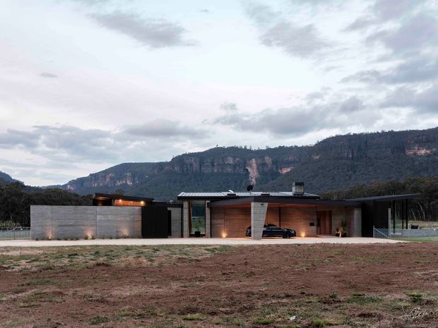 Single-storey contemporary residence set against a dramatic wooded escarpment. The low, horizontally composed building features dark metal roofing, concrete or rendered masonry walls, extensive glazing, warm timber-clad walls, and a covered carport or entry zone. The broad undeveloped foreground and surrounding mountain landscape emphasize the rural, elevated setting.