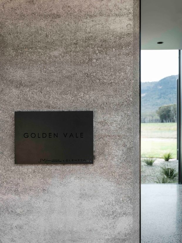 A close-up architectural view of a textured grey concrete or rendered wall with a dark rectangular “GOLDEN VALE” sign mounted to it. At right, a slim black-framed glazed opening reveals a bright landscaped rural setting and hills beyond. The interior has a smooth pale ceiling with a recessed downlight and a polished pale floor visible at the threshold.