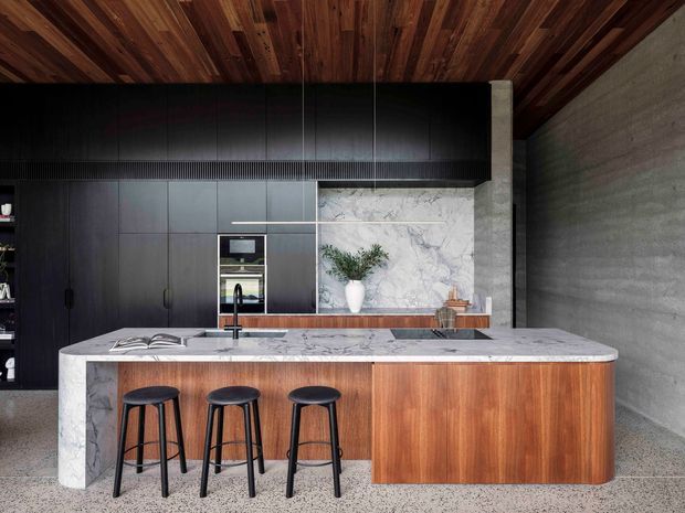A sophisticated contemporary kitchen centered on a large island with a white veined stone benchtop and warm timber-clad base. Three black stools sit along the island, facing dark matte cabinetry with integrated appliances. A marble-look splashback, concrete side wall, terrazzo-like floor, and rich timber ceiling create a refined, high-contrast material palette.