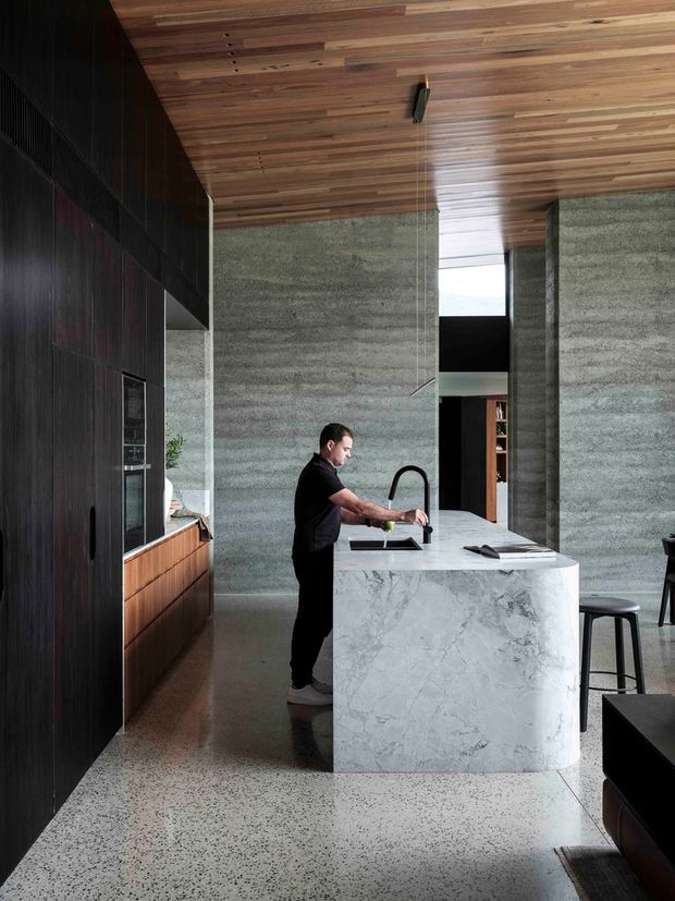 A contemporary kitchen viewed from within the open-plan living area, centred on a large sculptural white-veined stone island with an integrated sink and black gooseneck tap. Dark timber full-height cabinetry lines the left wall, with warm timber drawers and appliances built in. The room has textured grey-green concrete or plaster walls, a warm timber-lined ceiling, polished terrazzo flooring, high vertical openings, and black bar stools. The composition has a refined, material-focused architectural interior character.