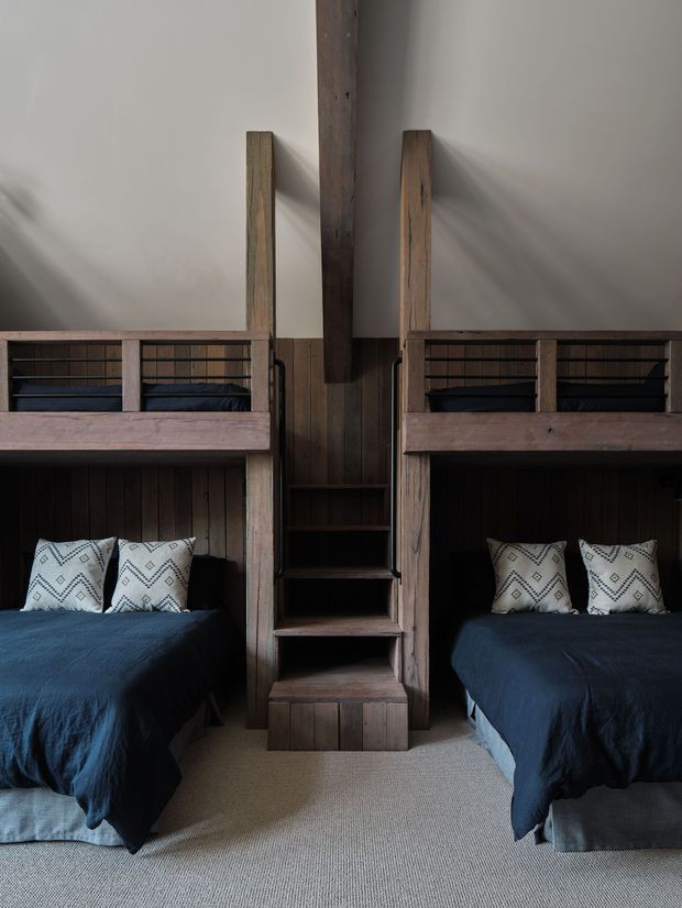 Symmetrical compact bedroom interior featuring two built-in bunk-style sleeping alcoves or beds on either side of a central timber staircase and shelving unit. The room has dark stained vertical timber panelling and joinery, deep navy bedding, patterned white cushions, off-white walls, and a light neutral carpet. The integrated timber construction and paired sleeping spaces create a cozy, efficient cabin-like arrangement.