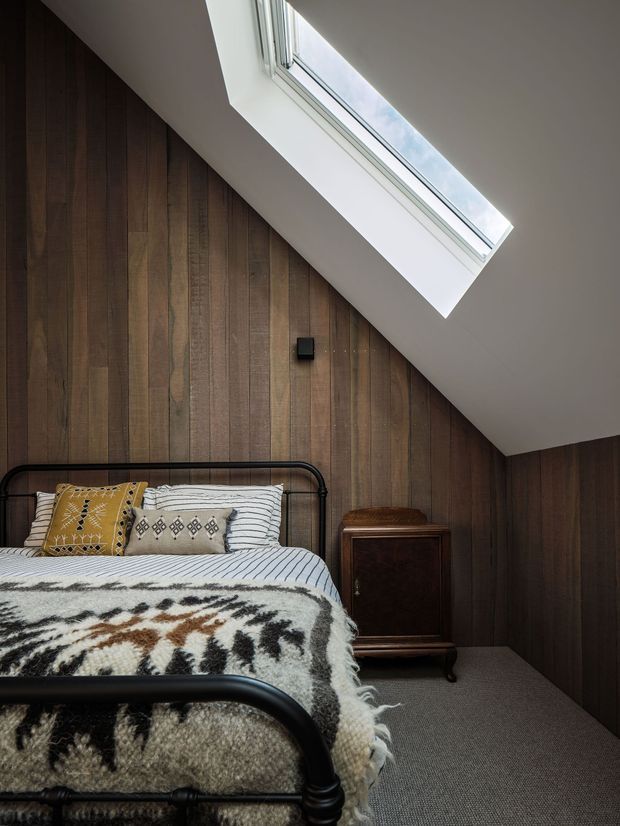 A compact attic bedroom with a black metal bed, patterned bedding, a vintage dark timber bedside cabinet, and full-height dark timber wall panelling beneath a sloping ceiling. A large roof skylight brings in natural light, while grey carpet covers the floor. The room has a warm, rustic and slightly eclectic character.