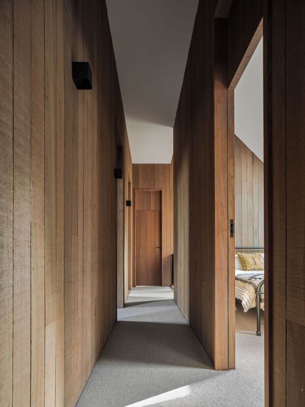 A narrow, atmospheric interior hallway lined with vertical warm-toned timber wall panelling and matching timber door frames. The corridor has a grey carpeted floor, a simple light ceiling, and small black wall-mounted light fittings. An open doorway on the right provides a partial view into a bedroom with a bed and patterned bedding. The composition emphasizes the long perspective, recessed door, and strong contrast between dark timber surfaces and daylight.