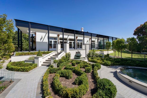 A low, elongated modern residence viewed from a landscaped garden, with extensive floor-to-ceiling glazing, slender dark window frames, pale rendered walls, and a flat or very low-pitched roof. Formal clipped hedges, geometric garden beds, paved paths, steps, and a curved pool edge define the foreground and side garden.