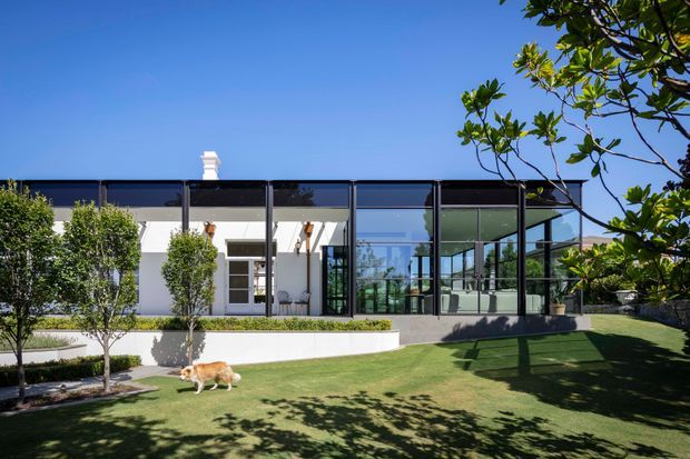 Exterior view of a low, modern residential building with a flat roof, white rendered walls, extensive black-framed floor-to-ceiling glazing, and a glazed pavilion-like living area. The house opens onto a manicured lawn with raised white garden edging and clipped trees, while mature foliage frames the right side of the image.