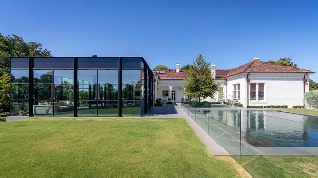 Exterior view of a refined traditional white villa with terracotta tiled roofs, connected to a starkly modern black-framed glazed pavilion or extension. The property features expansive lawns, a glass pool fence, and a long rectangular swimming pool, creating a clear contrast between historic-style architecture and contemporary addition.