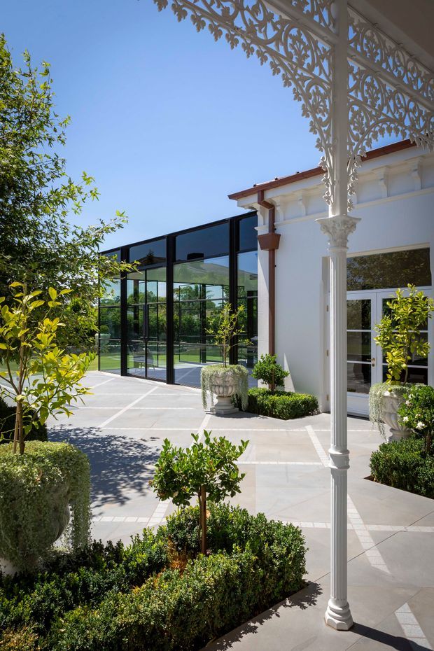 Sunlit formal courtyard beside a white heritage-style building, framed by ornate white decorative metalwork and columns. Pale large-format paving surrounds manicured box hedges, clipped topiary, and potted citrus trees. A black-framed glazed pavilion or conservatory extends across the rear, with a glimpse of a sports court beyond.