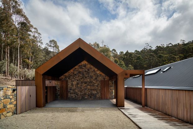 Exterior view of a contemporary gabled carport or covered entry structure set within a wooded landscape. The structure features a dark metal pitched roof, substantial warm timber framing, a rustic natural-stone rear wall, timber privacy fencing, a gravel driveway, and a narrow timber walkway. A connected building with roof skylights is visible to the right.