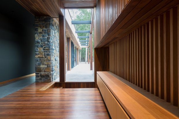 A narrow, refined timber-lined interior passage or entry space with a rich dark wood floor, vertical timber wall panelling, and integrated bench or storage joinery along the right side. A rough natural-stone column contrasts with the warm wood and dark painted wall on the left. Floor-to-ceiling glazing and a glazed overhead canopy lead toward a bright exterior courtyard or walkway with trees beyond, creating a strong indoor-outdoor connection.