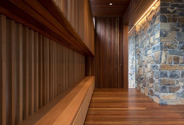 A narrow, warmly lit architectural passage or entry hall featuring extensive dark timber wall panelling, a built-in timber bench and deep shelving along the left side, a timber-lined ceiling, dark timber flooring, and a prominent rough stone feature wall on the right. Recessed and concealed linear lighting accentuate the rich natural materials and strong linear perspective.