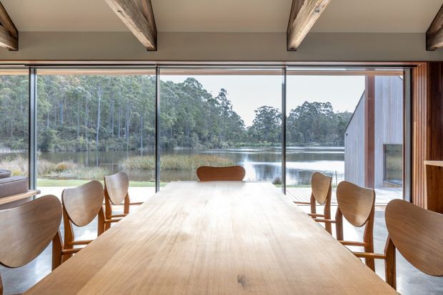A warm, timber-lined dining room centred on a long solid-wood dining table and sculptural wooden chairs. Full-height glazing frames a tranquil lake and forest landscape, while exposed timber ceiling beams and vertical timber wall panelling add rustic character. The room is bright, open, and strongly connected to the surrounding natural setting.