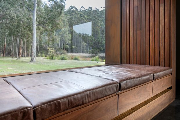 Interior architectural detail featuring a built-in window seat with brown leather cushions and warm timber drawer cabinetry. Vertical timber wall panelling frames a large glazed opening looking onto a wooded landscape, creating a warm, nature-connected retreat.