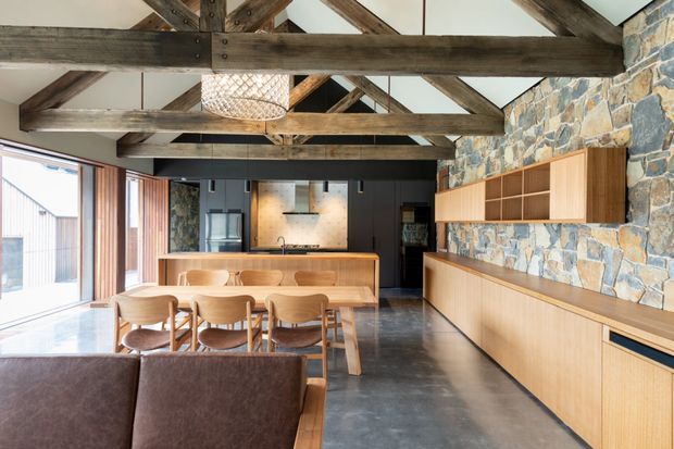 Open-plan kitchen and dining area in a rustic contemporary interior, featuring exposed dark timber roof trusses, a rough natural stone feature wall, polished concrete flooring, extensive timber cabinetry, a kitchen island, and a large dining table. Full-height glazing brings in abundant daylight, while a woven pendant light and warm timber furnishings add softness.