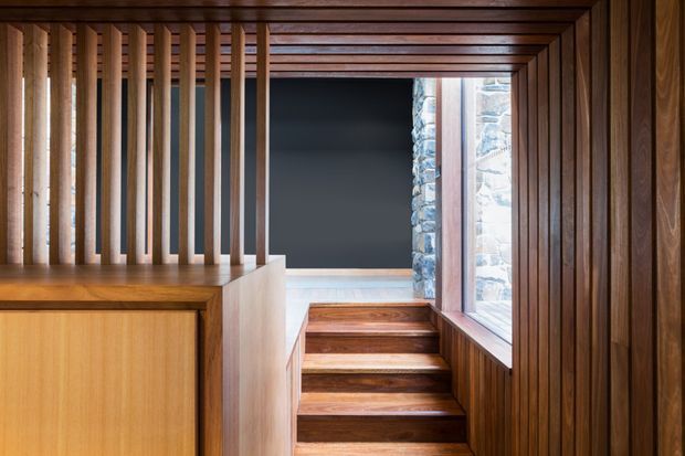 A richly timber-lined interior passage with a short flight of timber stairs, vertical timber slats forming a screen over built-in cabinetry on the left, and a large window on the right beside a textured natural-stone wall visible outside. The space has strong linear detailing, warm wood tones, and a dark opening or wall at the stair landing.