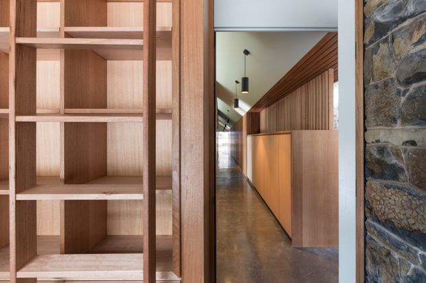 Interior architectural detail showing a custom timber built-in shelving unit in the foreground, opening onto a long polished concrete-floored hallway or reception-like space. The corridor features extensive vertical timber wall panelling, warm timber cabinetry, black pendant lighting, and a rough natural stone wall at the right edge. The composition has a refined contemporary character with strong linear detailing and warm natural materials.