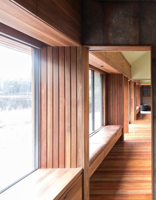 A narrow, sunlit interior corridor lined with rich vertical timber panelling and integrated timber window seats. Full-height glazing runs along the left side, while a large wall mirror reflects the continuous hallway, warm timber ceiling and flooring, and built-in bench seating. Dark textured stone or concrete-like wall panels frame the mirror and contrast with the wood. The space has a refined, warm, minimalist architectural character.