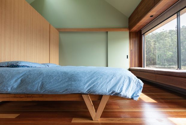 A warm, timber-lined bedroom with a low platform bed dressed in light blue bedding. The room features rich dark timber flooring, vertical timber wall panelling and built-in timber cabinetry around a large window seat. Pale green walls and ceiling create a calm, natural atmosphere, while the broad window looks onto a wooded landscape.