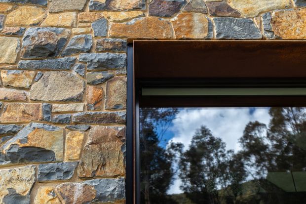Close-up architectural detail of a dark timber-framed window set into an irregular natural stone masonry wall. The window glass reflects trees and sky, while the stonework combines warm ochre, tan and grey tones with visibly varied block shapes and mortar joints.