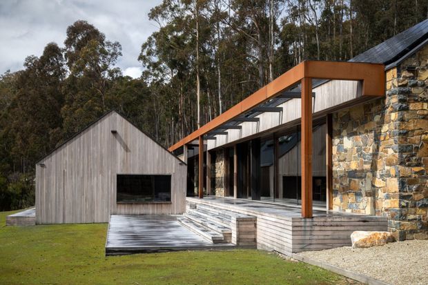 Exterior view of a contemporary multi-volume home set in a wooded landscape. The building combines a dark pitched metal roof, extensive glazing, warm orange-brown structural framing, weathered vertical timber cladding, and a prominent rough stone masonry wall. Broad timber decking and steps connect the volumes to the lawn.