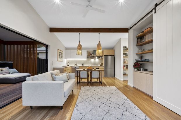 Open-plan interior showing a living room flowing into a compact kitchen and dining area. The room has white walls and ceiling, warm timber flooring, a large exposed timber beam, pale upholstered sofas, built-in white cabinetry with open timber shelving, a stainless-steel refrigerator, pendant lights, ceiling fans, and a patterned area rug. A bedroom is visible through a wide opening on the left, separated by dark vertical timber screening and glazing.