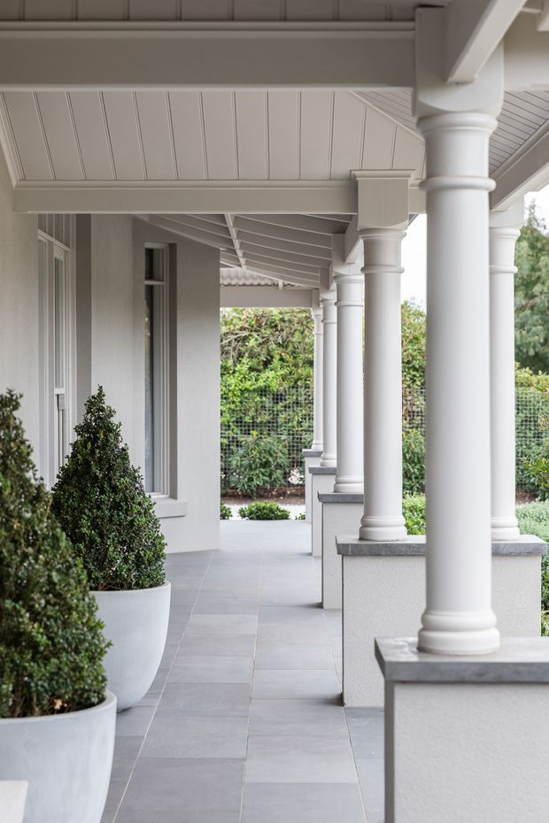 A covered exterior veranda or colonnaded walkway with repeated white classical columns, decorative capitals and bases, a paneled soffit, grey tiled flooring, and light rendered walls. Large white planters with clipped shrubs line the edge, with landscaped greenery visible beyond.